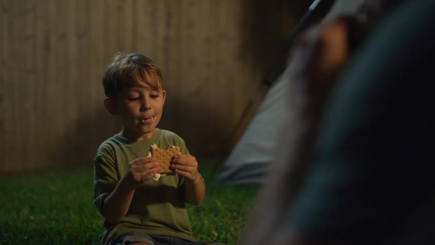 A father and young son eating smores while camping in a backyard