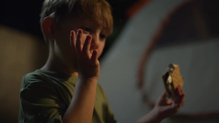 A young boy eats a smores outside at night beside a tent