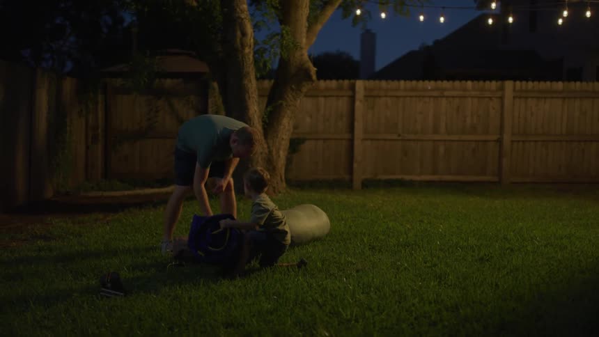 A father and son carry camping equipment into the backyard of a house and prepare to camp