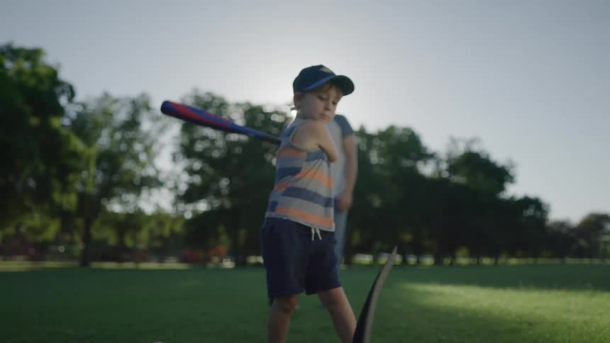 A boy trying to hit a baseball with a baseball bat on a t with a man trees the sky and clouds in the background