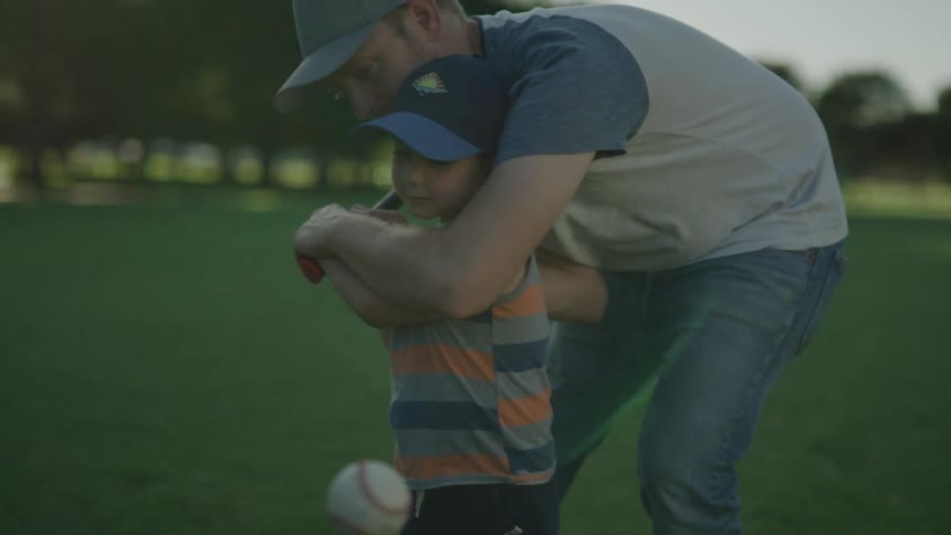 A father teaching his young son how to swing a baseball bat outside on a summer day