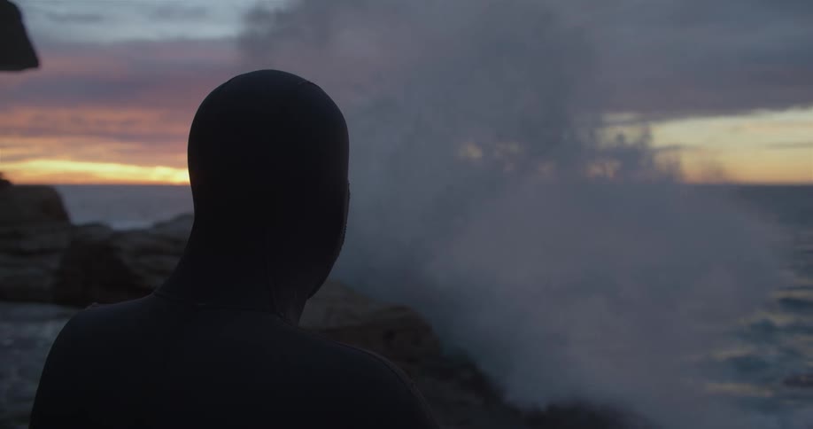 A silhouette of a person with a wetsuit on a rocky coast with the ocean sky and clouds in the background