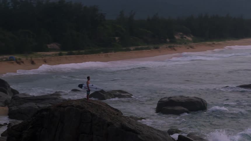 Young man standing over a rock near the ocean holding a surfboard at sunset