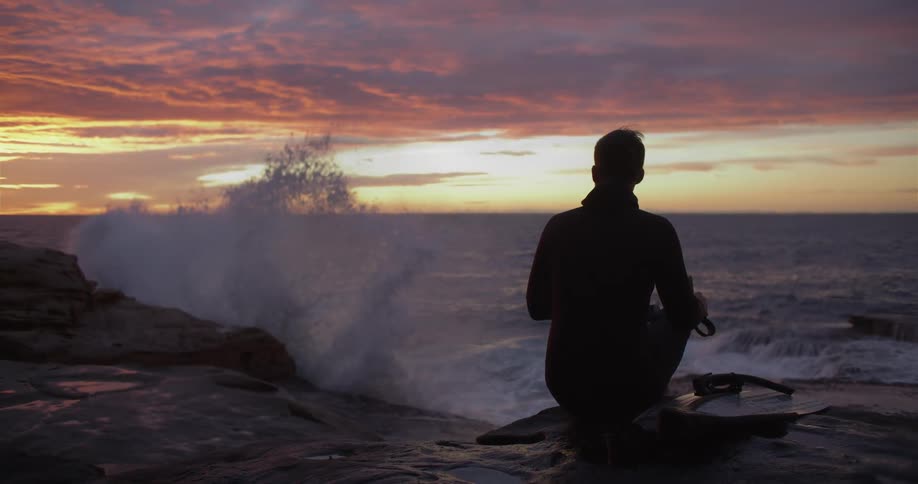 Silhouette of a man sitting on a beach watching the sunset as waves splash against the rocks