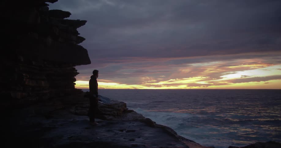 Man stands on the rocks and looks out at the ocean at dusk
