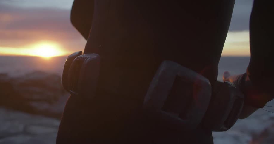 A person in a wetsuit strapping on a belt while standing on a beach at sunset