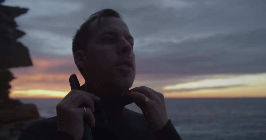 A diver sitting on the coast checks his mask and pulls the hood of his wet suit over his head as he prepares to dive at sunset