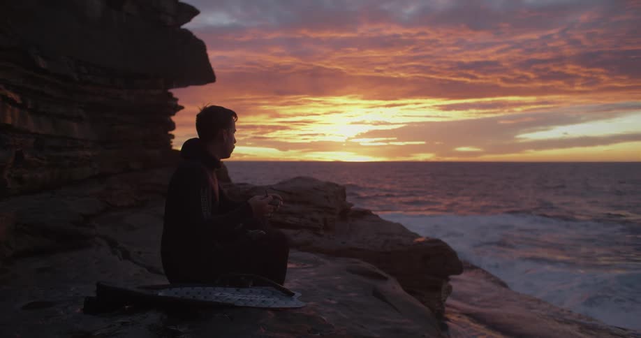 A man sitting outside on rocks near the ocean at a sunset