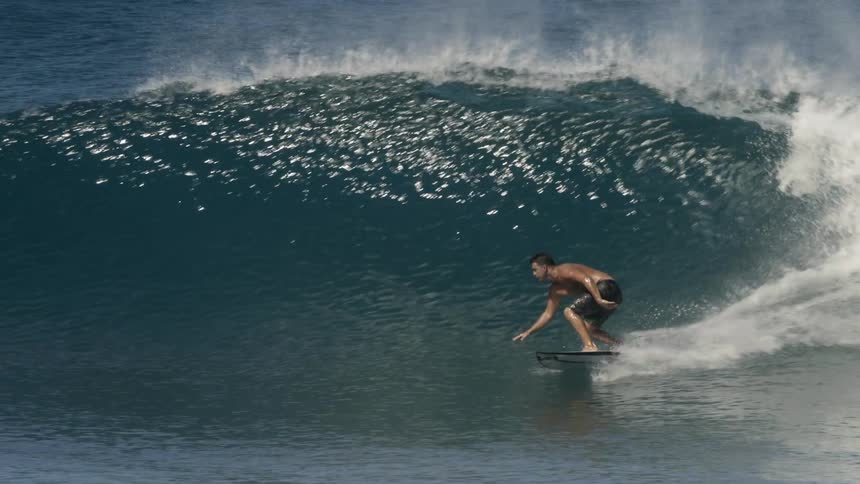 Surfer paddles near others before standing on surfboard to ride wave over reflecting water
