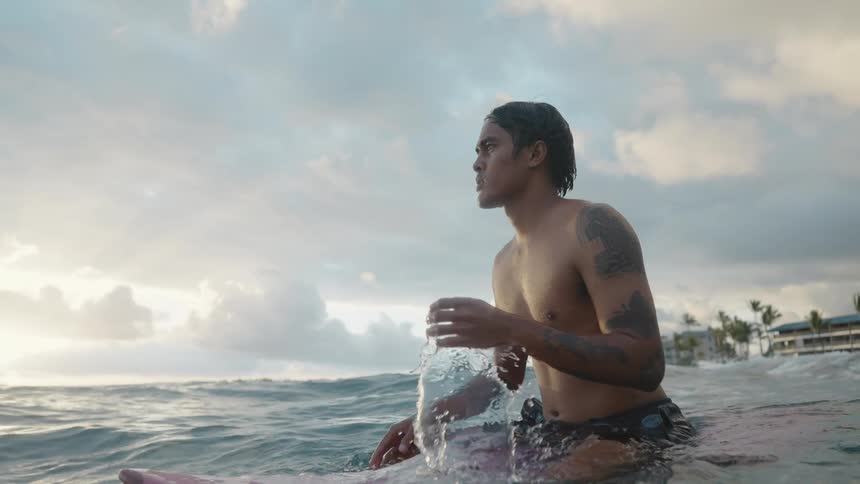 A man on a surfboard in the ocean with buildings trees the sky and clouds in the background