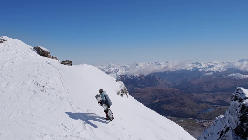 A snowboarder walks up the side of a tall mountain with a snowboard