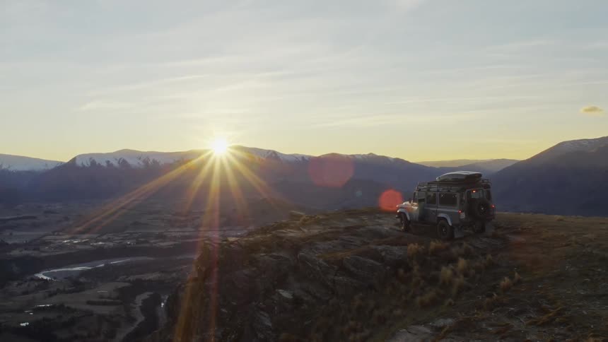Sun shines from just over snow topped mountain as man parks car on promontory overlooking valley before leaving car to walk to edge