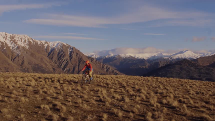 Man bicycles across slightly slopping ground with mountains capped by snow in the background
