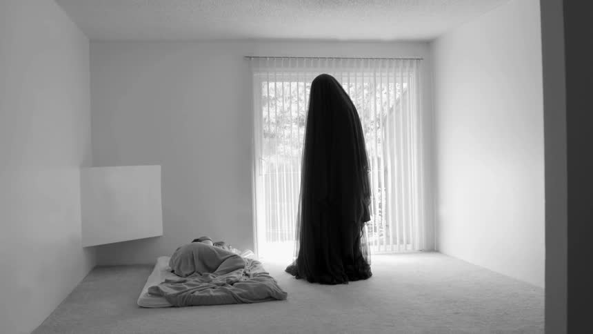 Cloaked figure standing over a sleeping person in a white room