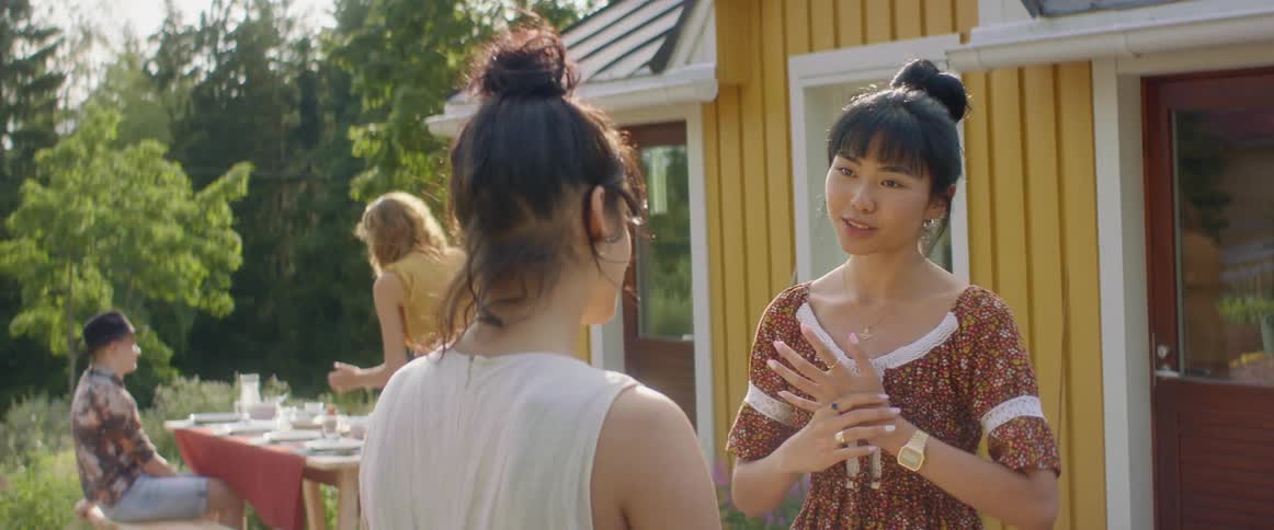 A pair of young women talk with one another while standing outside of a house during the day