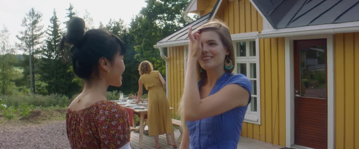 Friend greet each other with a hug as other friends congregate around picnic table with woods and field in backgrond