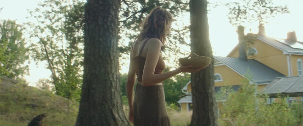 A woman holding a basket stands and walks away from hens toward her house