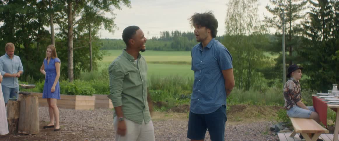 A group of people stand outside at a gathering and one man ruffles another mans hair in fun