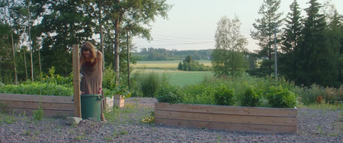A woman uses a watering pail to water plants in a backyard garden
