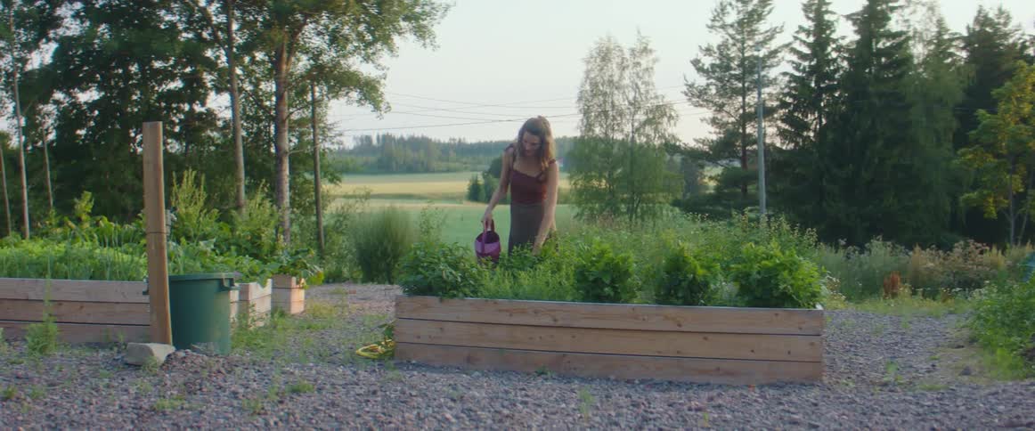 A woman uses a watering can to water vegetable plants in a raised garden box