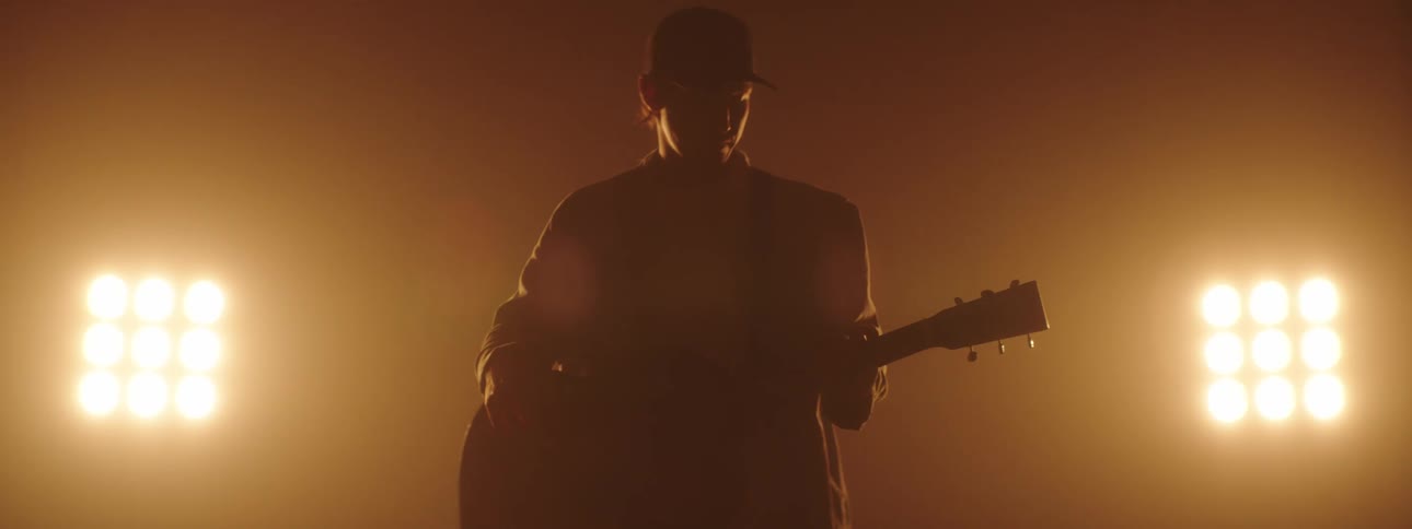 Bright lights shine on a man wearing a baseball cap and playing a guitar
