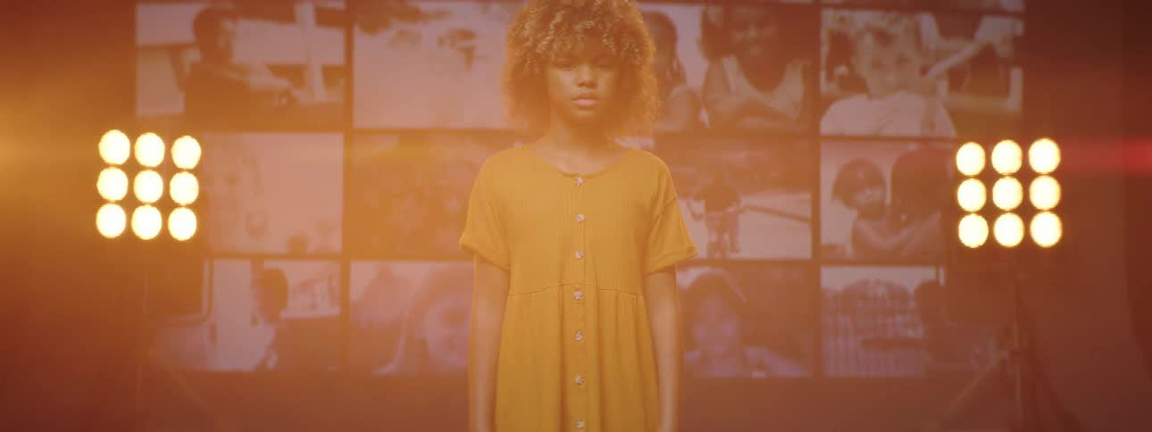 A girl stares into the camera while standing before stage lights and a video wall on stage
