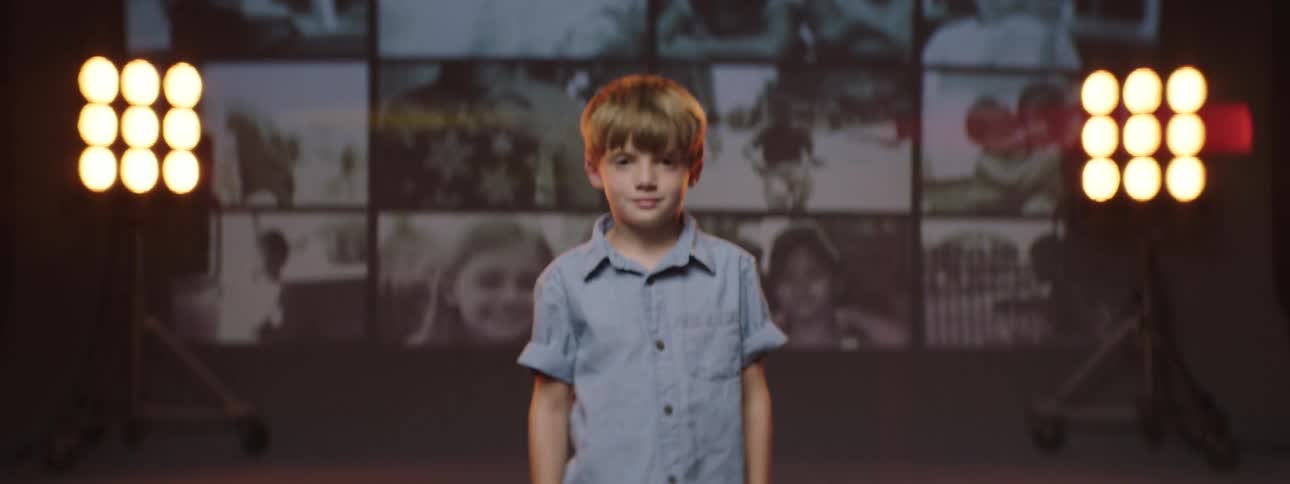 A boy standing and staring on a stage with spotlights pictures and a wall in the background
