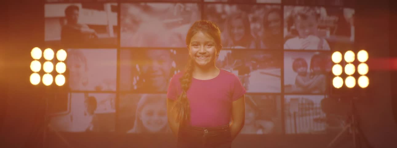 A girl standing and staring on a stage with spotlights a screen and a wall in the background