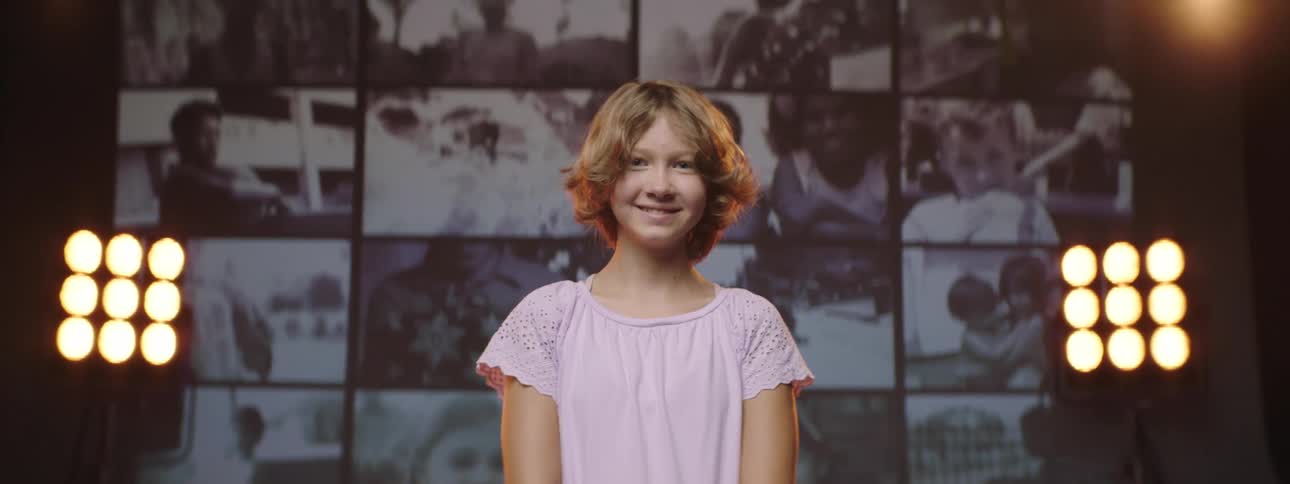 Teenager with short hair stands in front of a wall of monitors and smiles
