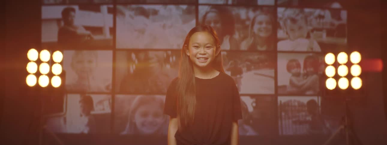 A girl smiles as she poses in front of a dozen screens playing videos of children