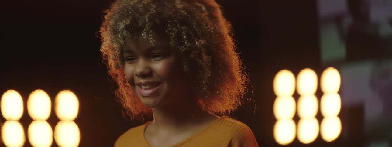 A girl standing and smiling on a stage with spotlights a screen and a wall in the background