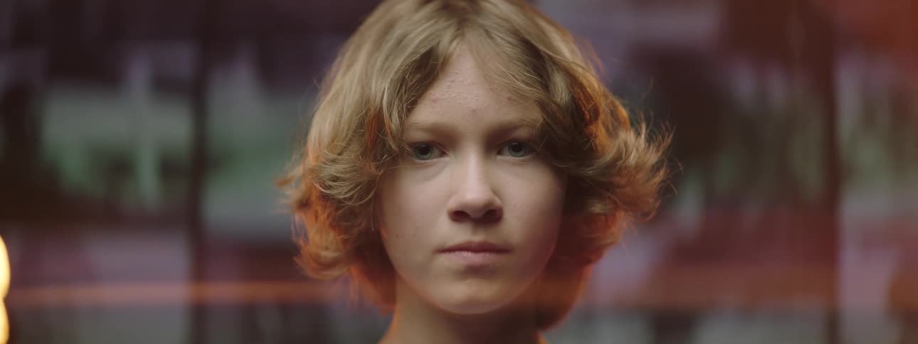 Blonde boy with long hair faces the camera with eye contact in front of a giant screen