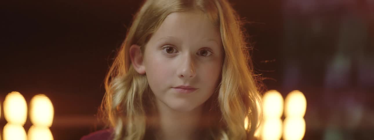A girl standing and staring on a stage with spotlights a screen and a wall in the background