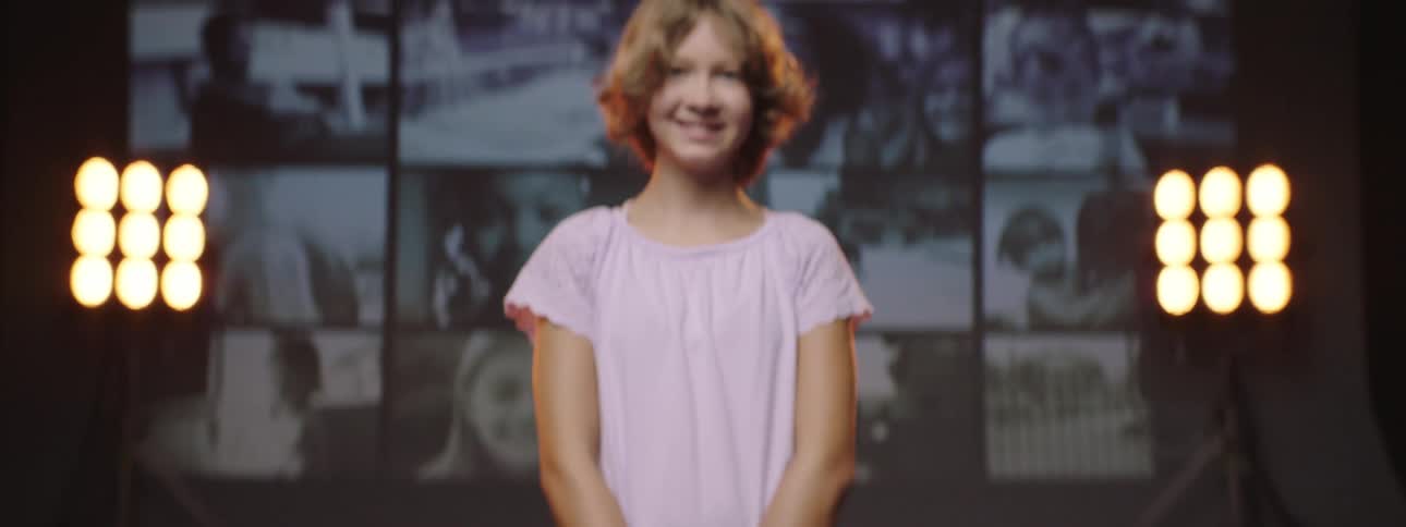 A young girl smiles as she stands in front of a screen playing multiple videos of other children