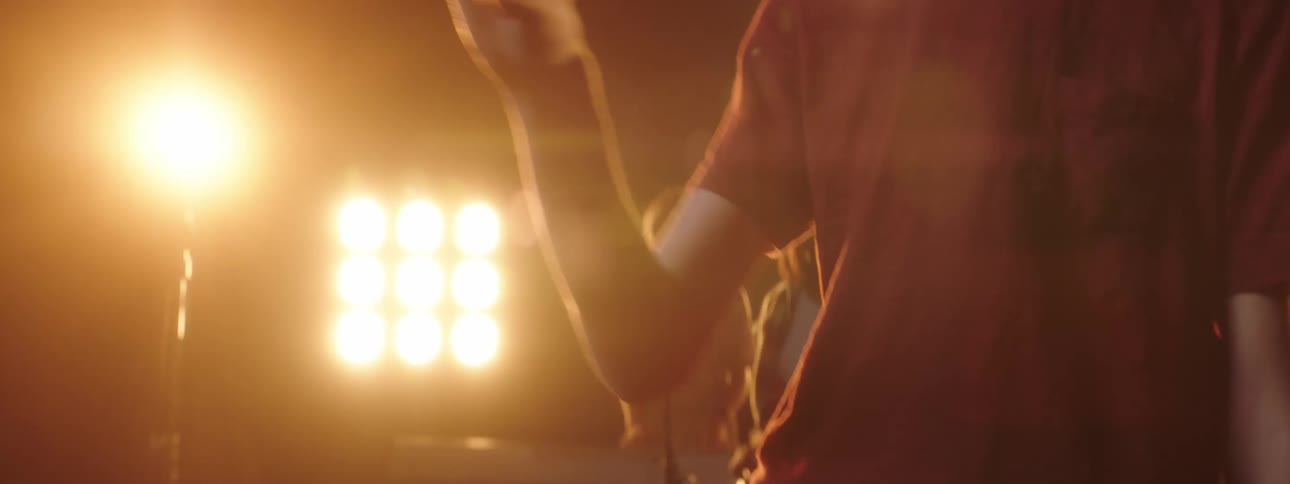 A man on stage twirl in a drumstick with people spotlights and a screen in the background