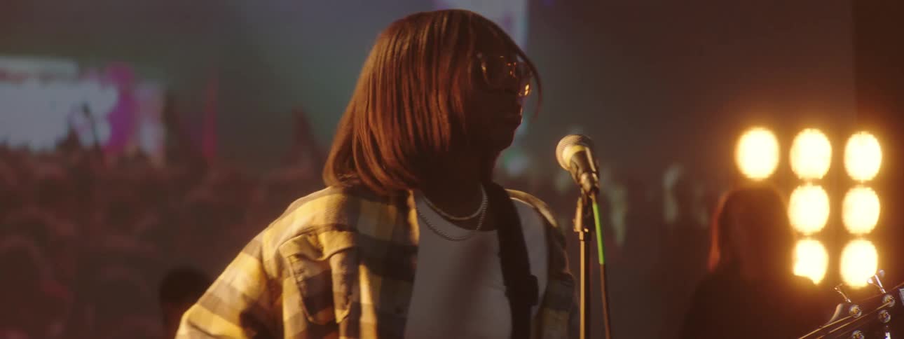 A girl on stage playing a guitar with people a spotlight and a screen in the background