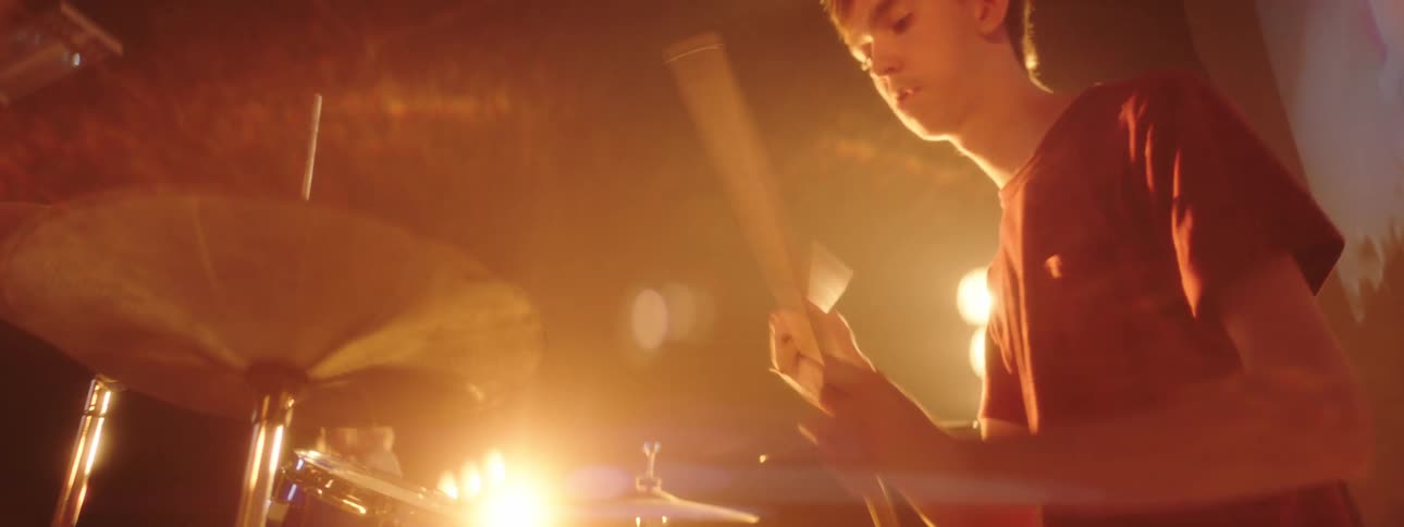 A man on a stage playing drums with drumsticks with a wall a screen and spotlights in the background