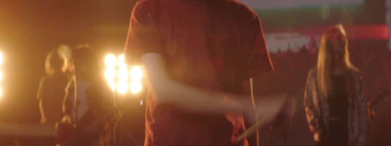 A teenager twirls drumsticks as a band stands behind him