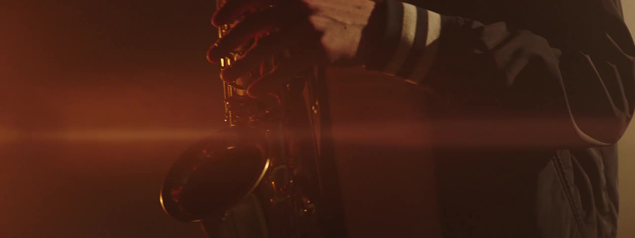 Man wearing a black jacket plays the saxophone as bright lights shine behind him