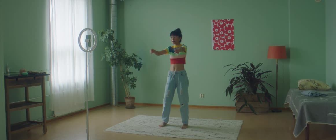 A young woman dances in a bedroom while recording herself with a smartphone on a stand