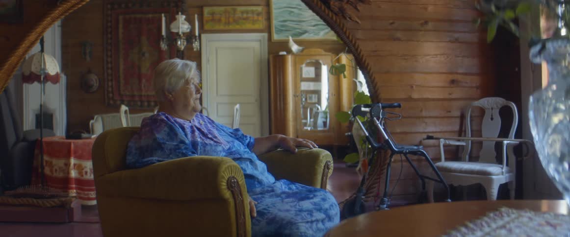 Elderly woman with grey hair and blue dress sits in brown armchair and looks around in the living room
