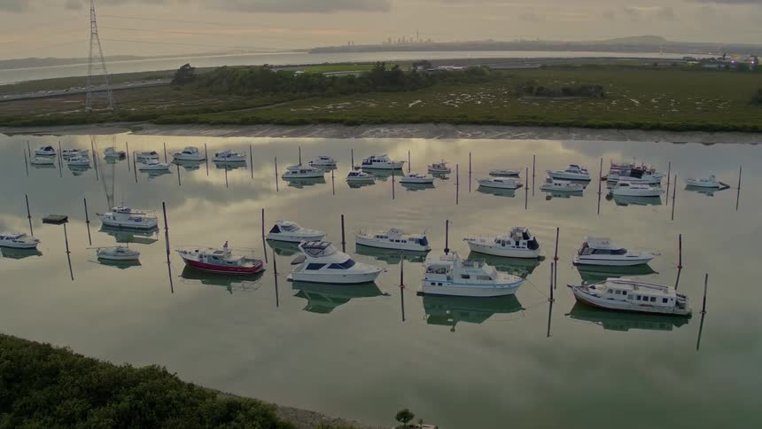 Boats sits moored on reflective water as traffic flows along highway near misty shore with skyline in far distance