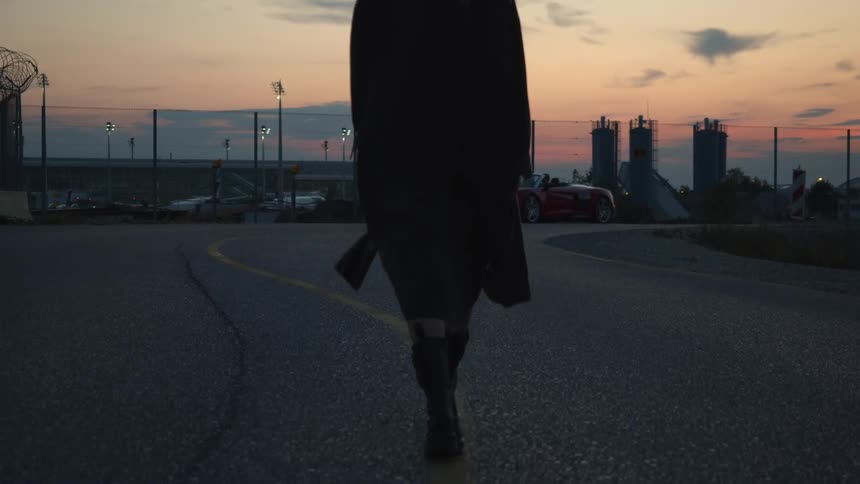 Woman wearing a long coat walks on the middle of the road as a convertible drives towards her