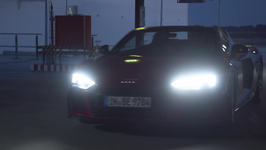 People drive across building parking lot in sports car with top down at night