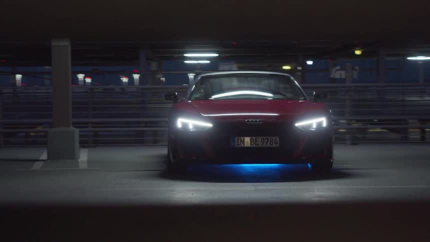 A red convertible sits in a parking garage with its headlights on