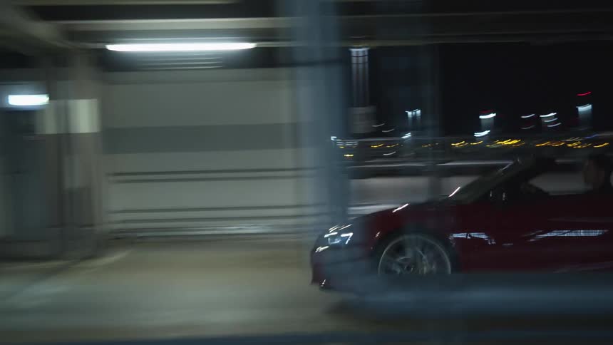 Person drives read convertible with top down along street with bright urban lights in background
