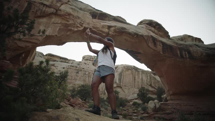 A young woman holds her phone taking a photo as she hikes through a canyon