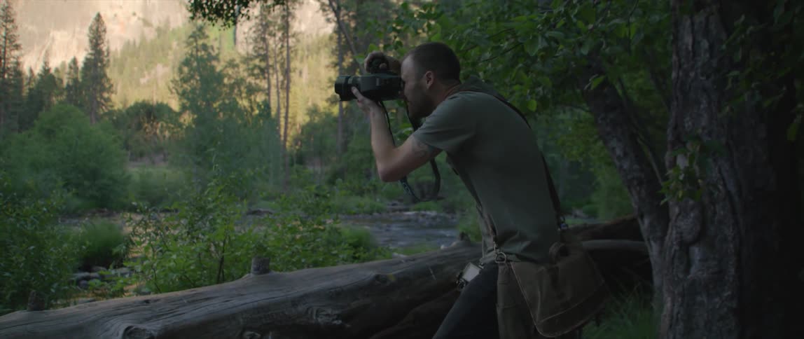 A man holds a professional camera to his face taking photos in a rural woods area