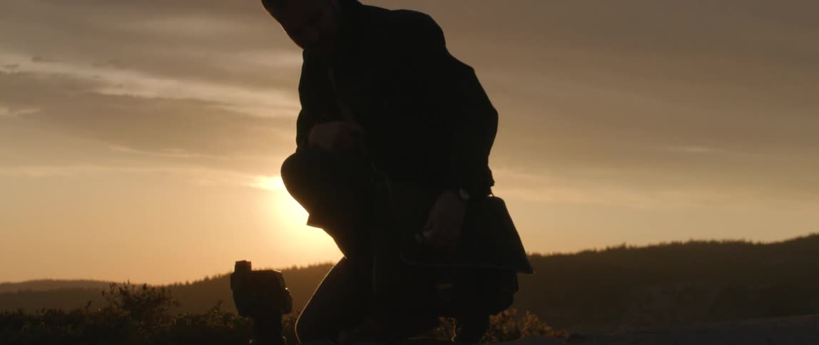A man kneels on the ground in a field while adjusting his camera