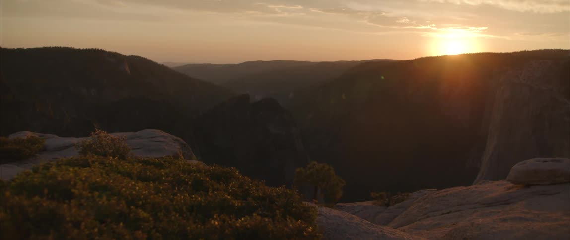 The sun shines brightly just above the horizon over a scenic canyon that is covered in vegetation